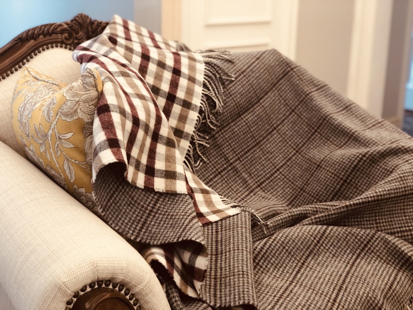 A plush, ornately carved sofa is draped with a patterned, brown and white checked blanket. A soft, floral-patterned pillow in gold hues complements the luxury of the setting.