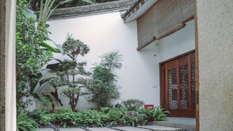 A serene courtyard features lush green foliage, with various plants and trees arranged harmoniously beside a white wall. A traditional wooden door with intricate carvings complements the natural elements. Stone pathways and a small red fire extinguisher add functional details.