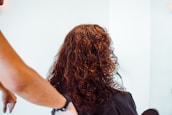A person with curly brown hair is being attended to, possibly in a salon environment. The arm of another individual is visible, suggesting hair styling or cutting.