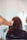 A person with brown, curly hair is seated in a chair, facing away. Another person, wearing a black bracelet, is holding scissors and preparing to cut the hair. Hairdressing tools are visible nearby, including a hair dryer attached to the wall. The environment suggests a salon setting with muted lighting and light-colored walls.