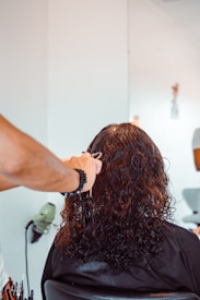 A person with brown, curly hair is seated in a chair, facing away. Another person, wearing a black bracelet, is holding scissors and preparing to cut the hair. Hairdressing tools are visible nearby, including a hair dryer attached to the wall. The environment suggests a salon setting with muted lighting and light-colored walls.