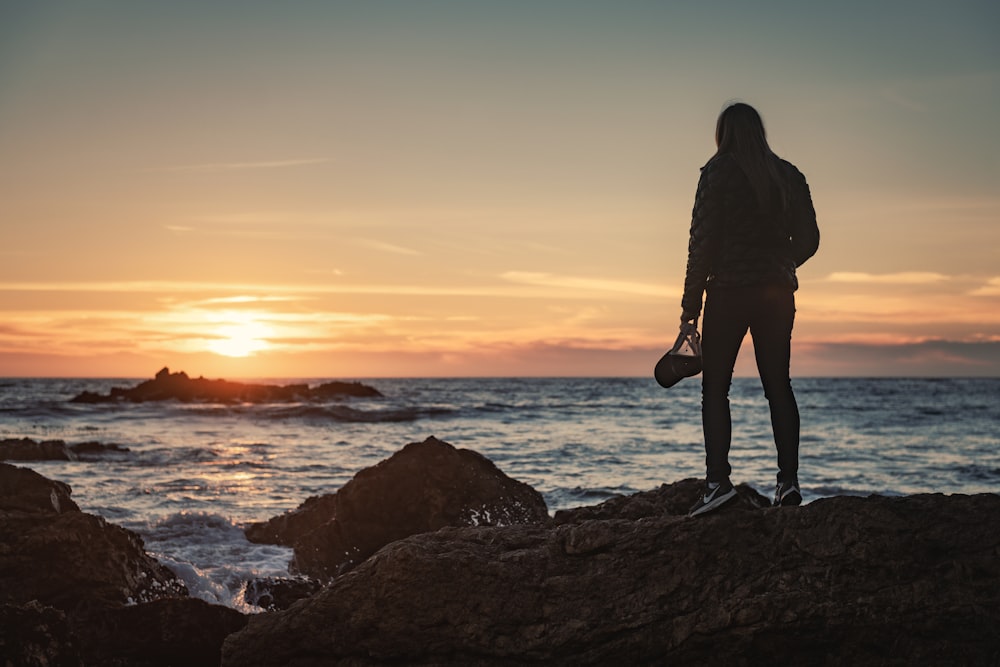 silhouette of man standing on rock formation near body of water during sunset