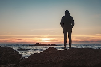 silhouette of man standing on rock formation near body of water during sunset