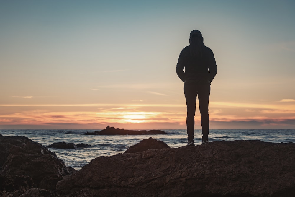 silhouette of man standing on rock formation near body of water during sunset