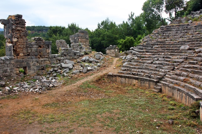 Ancient ruins are situated outdoors with remnants of stone structures and an amphitheater-like seating area carved from stone steps. The ground is a mix of dirt paths and patches of grass, while greenery, including trees and bushes, surrounds the area.