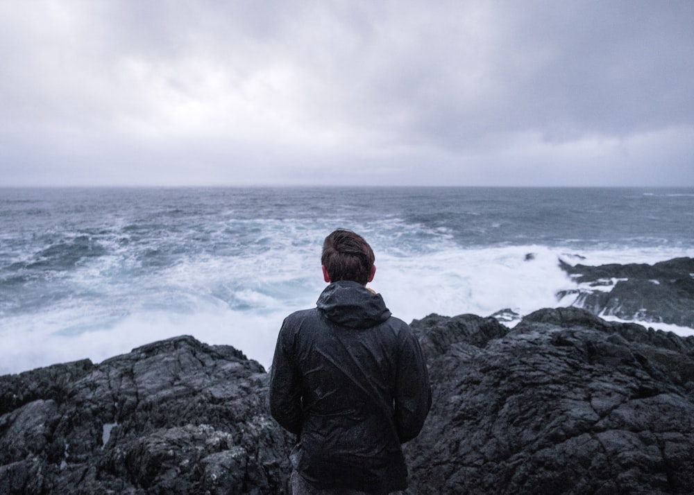 Mann in schwarzer Jacke sitzt tagsüber auf einer Felsformation in der Nähe des Meeres