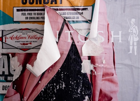 A collage of torn and layered posters reveals parts of text, graphics, and colors. Visible elements include portions of a pink jacket, a couple of text boxes, and a logo with 'Acklam Village' promoting street food and live music. The underlying layers and rips contribute to a textured appearance.