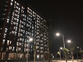 A tall residential building illuminated at night with numerous lit windows, indicating occupancy. Streetlights cast a warm glow on the trees and pavement below, while the sky is dark, creating a stark contrast with the building's lights.
