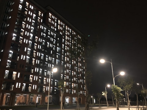 A tall residential building illuminated at night with numerous lit windows, indicating occupancy. Streetlights cast a warm glow on the trees and pavement below, while the sky is dark, creating a stark contrast with the building's lights.