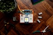 A small electronic project is assembled on a breadboard placed on a wooden surface. It includes wires, a microcontroller, and a display screen showing a humidity reading of 39% and a temperature of 25C. Various tools like wire cutters and digital components such as a seven-segment display are laid around it. A potted plant is visible in the top left corner.