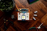 A small electronic project is assembled on a breadboard placed on a wooden surface. It includes wires, a microcontroller, and a display screen showing a humidity reading of 39% and a temperature of 25C. Various tools like wire cutters and digital components such as a seven-segment display are laid around it. A potted plant is visible in the top left corner.