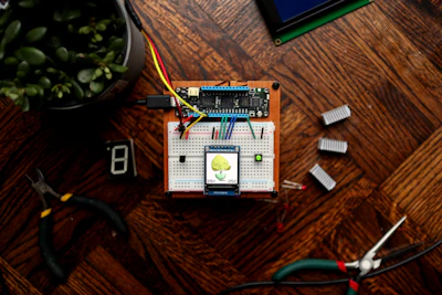 A small electronic project is assembled on a breadboard placed on a wooden surface. It includes wires, a microcontroller, and a display screen showing a humidity reading of 39% and a temperature of 25C. Various tools like wire cutters and digital components such as a seven-segment display are laid around it. A potted plant is visible in the top left corner.