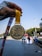 A hand holding a gold medal with the inscription 'Tata Mumbai Marathon 2020' is prominently displayed against a background of a marathon event, showcasing participants running on a city street. The medal's design features an embossed runner and text highlighting the marathon's completion distance.