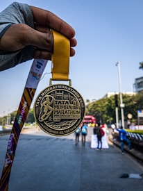 A hand holding a gold medal with the inscription 'Tata Mumbai Marathon 2020' is prominently displayed against a background of a marathon event, showcasing participants running on a city street. The medal's design features an embossed runner and text highlighting the marathon's completion distance.