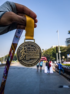 A hand holding a gold medal with the inscription 'Tata Mumbai Marathon 2020' is prominently displayed against a background of a marathon event, showcasing participants running on a city street. The medal's design features an embossed runner and text highlighting the marathon's completion distance.