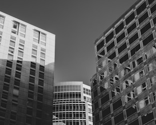 A black and white photograph captures several tall, modern office buildings. The structures have glass and steel facades with numerous windows reflecting light and shadows. The architectural lines are clean and geometric, with varying angles and perspectives.