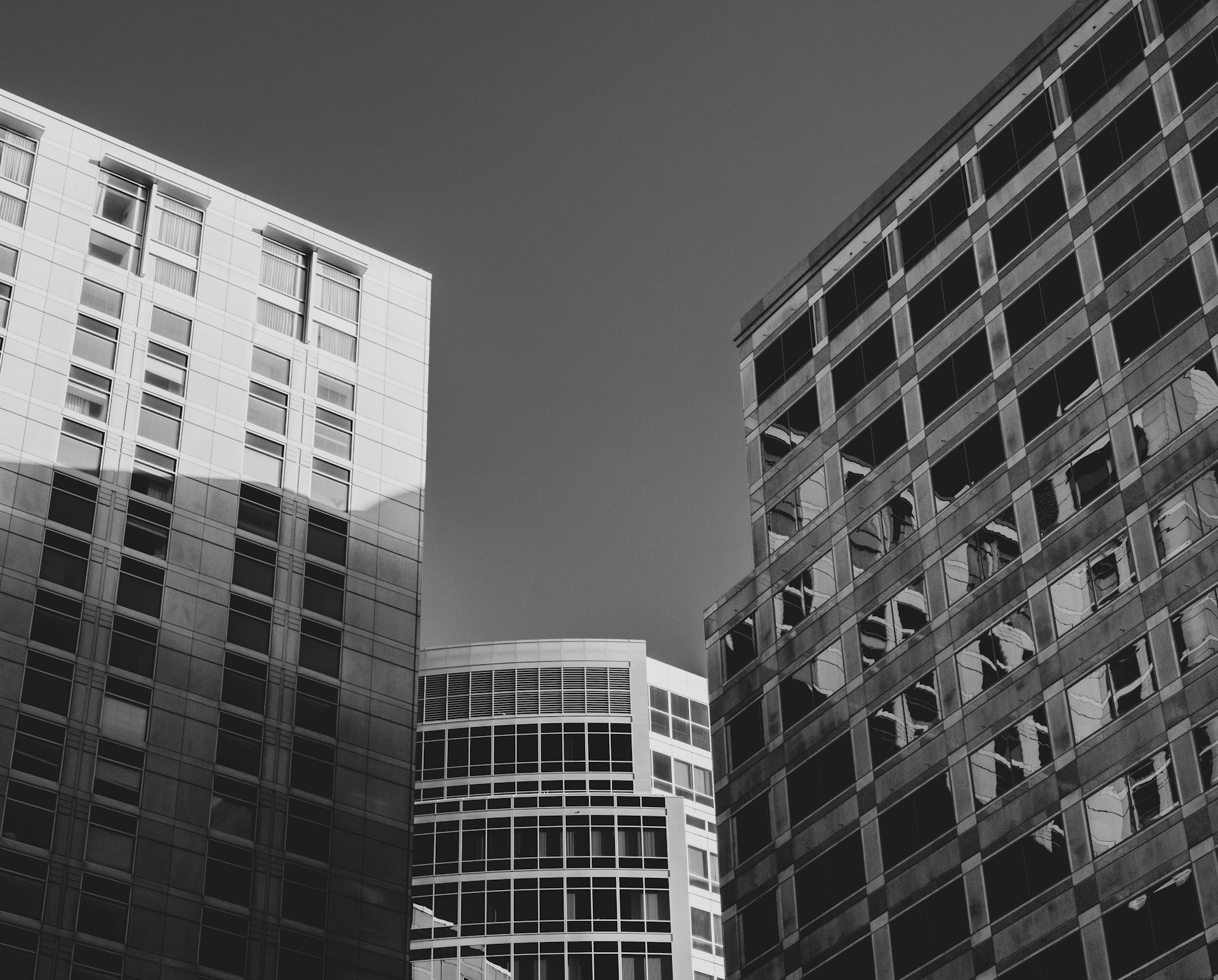 A black and white photograph captures several tall, modern office buildings. The structures have glass and steel facades with numerous windows reflecting light and shadows. The architectural lines are clean and geometric, with varying angles and perspectives.
