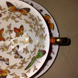 A close-up of a decorative teacup and saucer set embellished with colorful butterfly motifs and intricate floral patterns. The handle of the cup is metallic gold, adding a touch of elegance to the design.