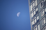 A partially illuminated moon is set against a clear blue sky alongside a modern glass building with numerous windows. The building occupies the right side of the image, providing a stark contrast to the natural element of the moon.