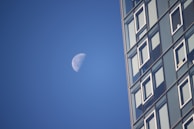A partially illuminated moon is set against a clear blue sky alongside a modern glass building with numerous windows. The building occupies the right side of the image, providing a stark contrast to the natural element of the moon.