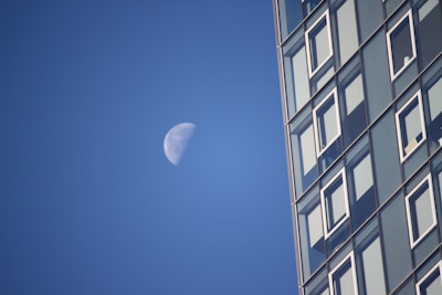 A partially illuminated moon is set against a clear blue sky alongside a modern glass building with numerous windows. The building occupies the right side of the image, providing a stark contrast to the natural element of the moon.