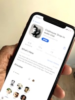 A hand holding a smartphone displaying the app page for Clubhouse: Drop-in audio chat on the screen. The app is available to open, with information such as user rating, age suitability, developer details, and a 'What's New' section visible. Several profile pictures are also displayed in a preview section.