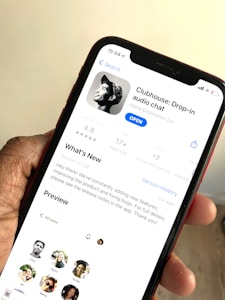 A hand holding a smartphone displaying the app page for Clubhouse: Drop-in audio chat on the screen. The app is available to open, with information such as user rating, age suitability, developer details, and a 'What's New' section visible. Several profile pictures are also displayed in a preview section.