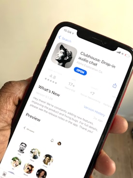 A hand holding a smartphone displaying the app page for Clubhouse: Drop-in audio chat on the screen. The app is available to open, with information such as user rating, age suitability, developer details, and a 'What's New' section visible. Several profile pictures are also displayed in a preview section.