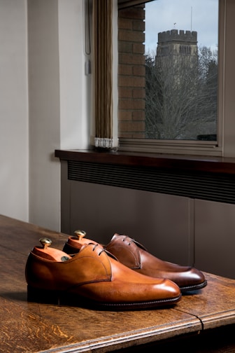 A pair of polished brown leather shoes is placed on a wooden table in an indoor setting. The shoes have a classic design with a lace-up front. In the background, there is a window with blinds partially drawn, offering a view of an outside stone tower surrounded by bare trees, suggesting the season might be winter or early spring.