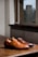 A pair of polished brown leather shoes is placed on a wooden table in an indoor setting. The shoes have a classic design with a lace-up front. In the background, there is a window with blinds partially drawn, offering a view of an outside stone tower surrounded by bare trees, suggesting the season might be winter or early spring.