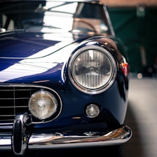 Close-up of a classic car's front, showcasing its round headlights, chrome bumper, and deep blue body. The shine on the metal and the rich color contribute to a vintage aesthetic.