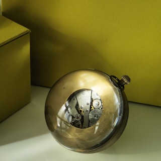 A spherical metallic object with visible internal gears and mechanical parts, set against a neutral olive green background. The object appears to be a dismantled or transparent clock or watch, showcasing its intricate mechanisms. The setting includes a corner where two walls meet, emphasizing minimalism.