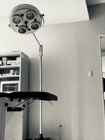 A medical examination room features an overhead surgical lamp and a mechanized adjustable examination table. The room has a cabinet filled with various medical supplies and tools, and the walls are bare with an electrical outlet visible near the floor.