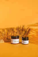Two jars labeled 'Pain Relief CBD Salve' are placed on an orange surface, accompanied by a shadow of a decorative palm leaf. The larger jar is on the left, while the smaller one is on the right. The backdrop is a gradient of warm yellow shades, conveying a natural and organic ambiance.