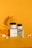 A composition of CBD salve packaging and jars is arranged artistically against a vibrant orange background. Shadows of a plant leaf are cast across the scene, adding texture and depth. Small porous stones are scattered around the products, enhancing the natural theme.