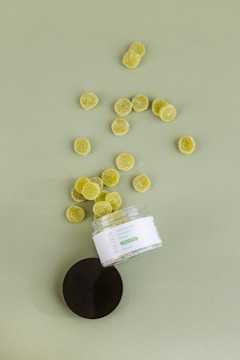 An open jar labeled 'Flavored CBD Gummies' with a number of round, green gummy candies spilled out onto a flat surface. The lid of the jar is lying nearby.