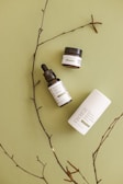 A minimalist arrangement featuring a dropper bottle, a jar, and a cylindrical container with skincare product labels on a light green surface. Delicate brown twigs are placed artistically around the products, enhancing the natural aesthetic.