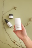 A hand holding a white container labeled 'Eczema Treatment CBD Ointment 500mg' against a soft green background. Nearby, there are two other skincare product containers, including a dropper bottle. A thin branch with small buds is also visible, adding a natural element to the composition.