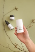 A hand holding a white container labeled 'Eczema Treatment CBD Ointment 500mg' against a soft green background. Nearby, there are two other skincare product containers, including a dropper bottle. A thin branch with small buds is also visible, adding a natural element to the composition.