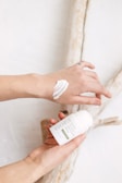 Hands are applying a white cream from a container labeled 'nora Eczema Hydro Cream' onto the skin. The background consists of light-colored wooden branches.