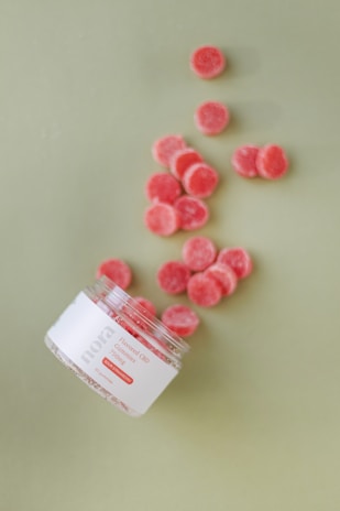 A tipped-over jar labeled 'nora Flavored CBD Gummies' scatters red gummies across a light green surface. The gummies are small, circular, and have a sugar coating.