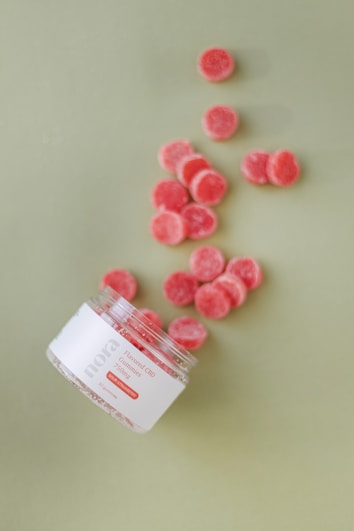 A tipped-over jar labeled 'nora Flavored CBD Gummies' scatters red gummies across a light green surface. The gummies are small, circular, and have a sugar coating.