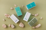 Several rectangular boxes with product labels, placed among natural coral stones on a green surface. The boxes have a minimalist design with colors like teal, light blue, white, and muted green.