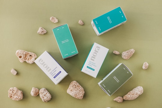 Several rectangular boxes with product labels, placed among natural coral stones on a green surface. The boxes have a minimalist design with colors like teal, light blue, white, and muted green.