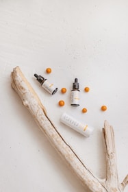 A minimalistic arrangement featuring skincare products, including one tall and one small bottle with dropper caps, along with several orange spheres. The objects are placed on a textured, light-colored surface accompanied by a piece of driftwood, adding a natural element to the composition.