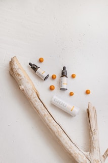 A minimalistic arrangement featuring skincare products, including one tall and one small bottle with dropper caps, along with several orange spheres. The objects are placed on a textured, light-colored surface accompanied by a piece of driftwood, adding a natural element to the composition.