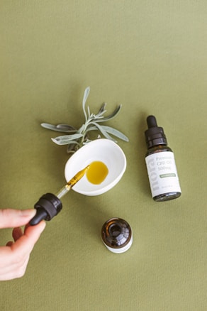 A dropper filled with golden liquid is being held above a small white dish, which contains some of the liquid. A bottle labeled as Premium CBD Oil 500mg lies on the olive-green surface beside the dish. A sprig of sage or similar herb is placed next to the dish, contributing to the natural aesthetic.