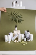 A collection of skincare products is artfully arranged on an olive green backdrop. Several cylindrical bottles and jars, some with labels, are prominently displayed. A large leaf is being held above the setup, adding a natural, organic element. Small rocks and green gummies are scattered around, enhancing the aesthetic presentation.
