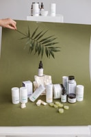 A collection of skincare products is artfully arranged on an olive green backdrop. Several cylindrical bottles and jars, some with labels, are prominently displayed. A large leaf is being held above the setup, adding a natural, organic element. Small rocks and green gummies are scattered around, enhancing the aesthetic presentation.