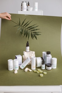 A collection of skincare products is artfully arranged on an olive green backdrop. Several cylindrical bottles and jars, some with labels, are prominently displayed. A large leaf is being held above the setup, adding a natural, organic element. Small rocks and green gummies are scattered around, enhancing the aesthetic presentation.